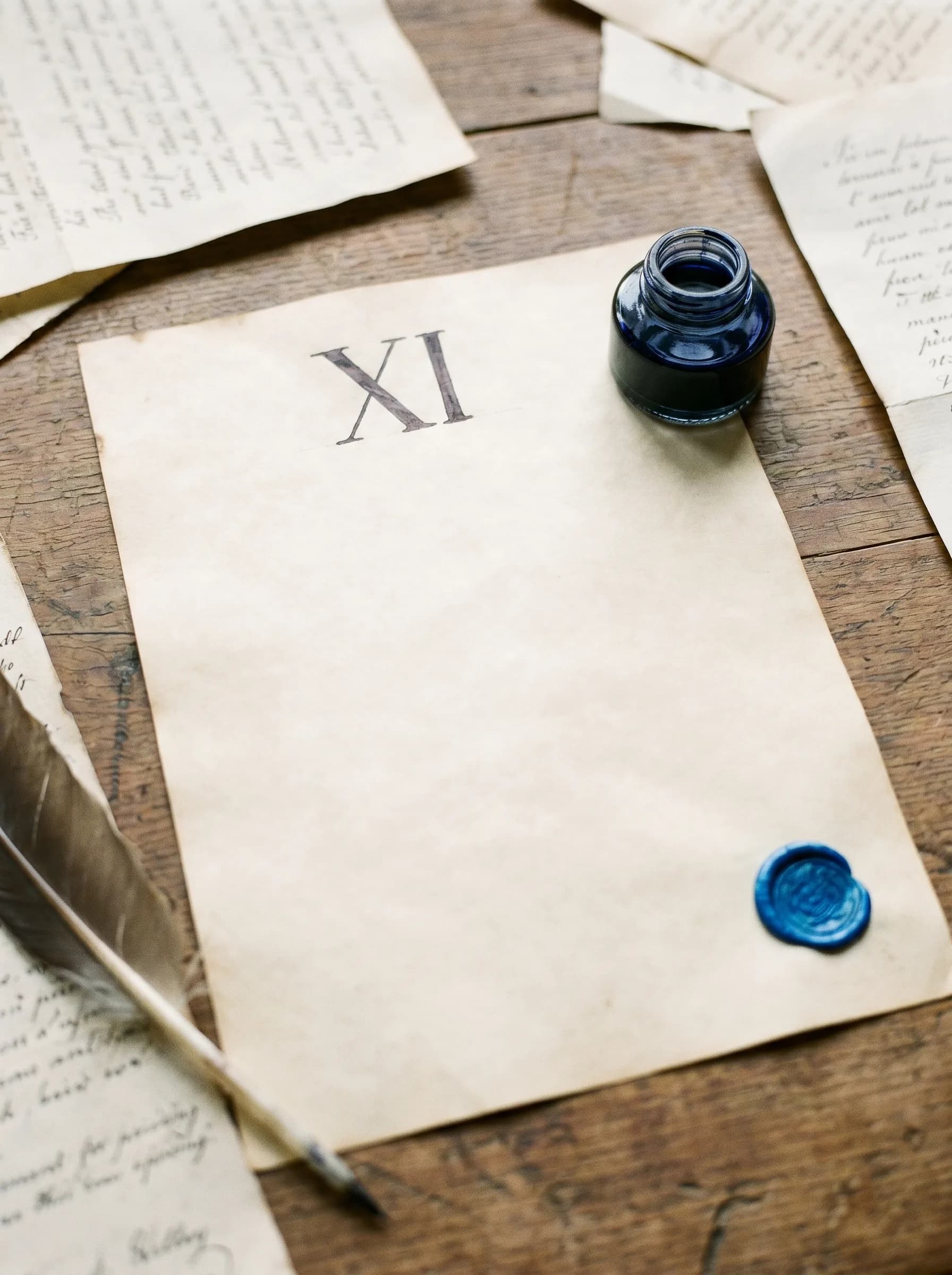 A sheet of aged cream paper marked with a single Roman numeral XI in navy ink, surrounded by other handwritten manuscript pages, a glass ink bottle and a feather quill resting on the desk.